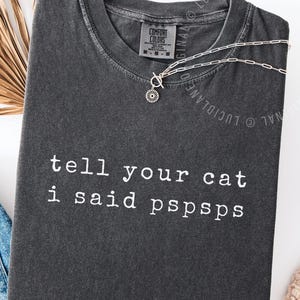Puede incluir: Camiseta gris oscuro con el texto blanco "tell your cat i said pspsps". Un collar plateado con un colgante de flores está colocado sobre la camiseta. La palabra "pepper" está impresa en la parte inferior.