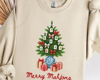 May include: Sand-colored sweatshirt with a festive design. A Christmas tree made of green branches is decorated with Mahjong tiles and red bows. Below, the words "Merry Mahjong" are written in red. Presents and a blue vase complete the design.