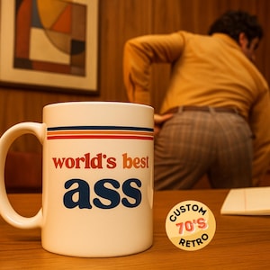 Puede incluir: Taza de cerámica blanca con el texto "world's best ass" en naranja y azul. La taza tiene un diseño retro con rayas naranjas, azules y marrones. Una pegatina "Custom 70's Retro" está sobre la mesa. El fondo incluye una persona y un cuadro enmarcado.