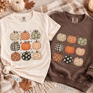 May include: An ivory t-shirt and an espresso brown sweatshirt, both featuring a grid graphic of nine pumpkins with various patterns including floral, plaid, polka dot, and heart designs. The garments have crew necklines and short or long sleeves.