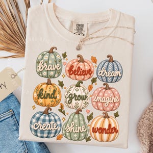May include: An ivory t-shirt featuring a graphic print of nine pumpkins in various colors and patterns. Each pumpkin displays a word in cursive script: brave, believe, dream, kind, grow, imagine, create, shine, and wonder. The shirt has a crew neckline.