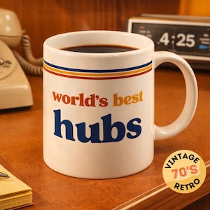 Puede incluir: Taza de cerámica blanca llena de café, con el texto "world's best hubs" en naranja y azul. La taza tiene un diseño retro con rayas y está sobre un escritorio de madera con un teléfono y un reloj vintage.