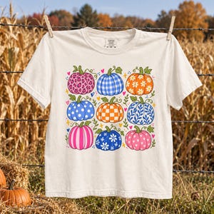 May include: A cream-coloured short-sleeve t-shirt featuring a graphic print of nine pumpkins arranged in a grid. The pumpkins display various patterns including leopard print, blue gingham, orange daisies, blue polka dots, orange checkers, and pink hearts.