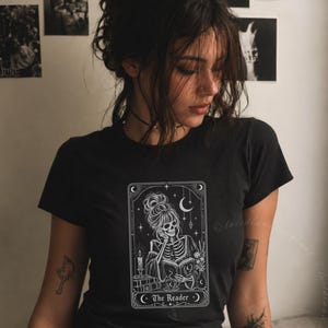Może przedstawiać: Czarny t-shirt z krótkim rękawem z białym nadrukiem graficznym przedstawiającym szkielet z kokiem czytający książkę. Wzór jest oprawiony jak karta tarota z elementami niebieskimi, świecą i kwiatami, zawierający napis The Reader na dole.