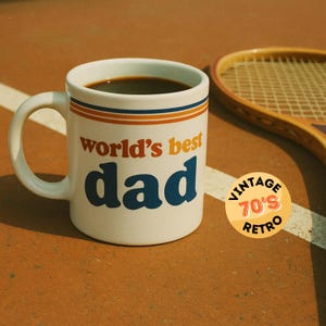 May include: White ceramic coffee mug with blue and orange stripes and the text "world's best dad". The mug is sitting on a brown surface with a tennis racket and a yellow circle with the text "Vintage 70's Retro" in the background.