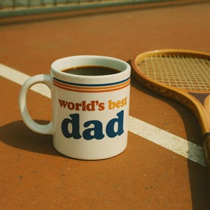 May include: White ceramic coffee mug with blue and orange stripes and the text "world's best dad". The mug is filled with coffee and is sitting on a brown surface next to a tennis racket.