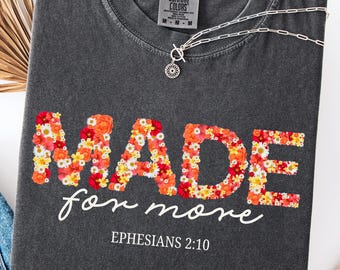 Op de afbeelding: Donkergrijs t-shirt met de woorden "MADE for more" in een bloemenontwerp. De tekst "Ephesians 2:10" staat eronder. Een zilveren ketting met een hanger ligt op het shirt.