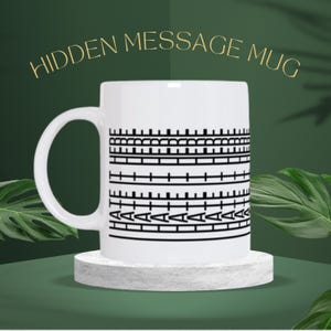 Op de afbeelding: Witte keramische mok met een zwart geometrisch patroon en de woorden "HIDDEN MESSAGE MUG" in gouden letters. De mok staat op een kleine, witte, vierkante sokkel.