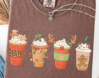 Op de afbeelding: Espresso-kleurig T-shirt met vier feestelijke koffiebekerontwerpen met vakantiethema's. De ontwerpen omvatten peperkoekmannetjes, rendiergeweien en decoratieve elementen. Het shirt heeft een ronde hals en een Comfort Colors-label.