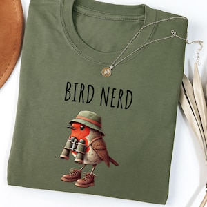 May include: An olive green t-shirt with the words "BIRD NERD" in black. A cartoon robin wearing a green hat, brown boots, and holding binoculars is printed below the text. A silver necklace with a pendant is draped on the shirt.