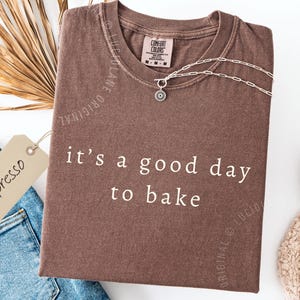 May include: A brown t-shirt with the phrase "it's a good day to bake" in white lettering. The shirt is folded and has a tag that says "espresso". A silver necklace and a pair of jeans are also in the image.