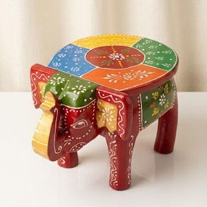 Puede incluir: Un pequeño taburete de madera pintado a mano con forma de elefante. El asiento presenta un diseño circular dividido en segmentos amarillos, verdes, azules y naranjas con motivos florales blancos. El cuerpo del elefante es rojo con detalles dorados y blancos.