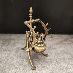 May include: A brass-colored, ornate incense burner with a tripod base and a handle featuring antler-like designs. The burner has a round body and a decorative top, with a spike extending upwards. The overall design is detailed and antique-inspired.