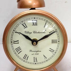 May include: A copper-toned alarm clock with Roman numeral markings on the face. The clock face reads "Village Clockworks Birmingham est. 1792".