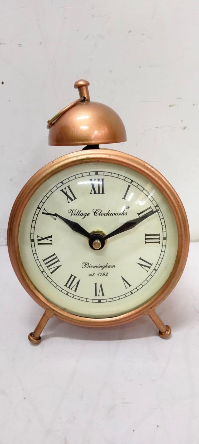 May include: A copper-toned alarm clock with Roman numeral markings on the face. The clock face reads "Village Clockworks Birmingham est. 1792".
