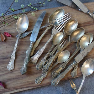 Op de afbeelding: Een assortiment antiek zilverwerk, waaronder lepels, vorken en messen, gerangschikt op een houten snijplank. Het zilverwerk heeft sierlijke handvatten en een aangeslagen zilveren afwerking. De opstelling bevat decoratieve takjes en bloemblaadjes.