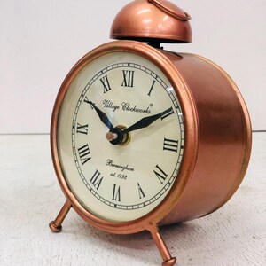 May include: A copper-colored alarm clock with Roman numeral markings on the face. The clock has a bell on top and three legs. The clock face reads "Village Clockworks Birmingham est. 1792".