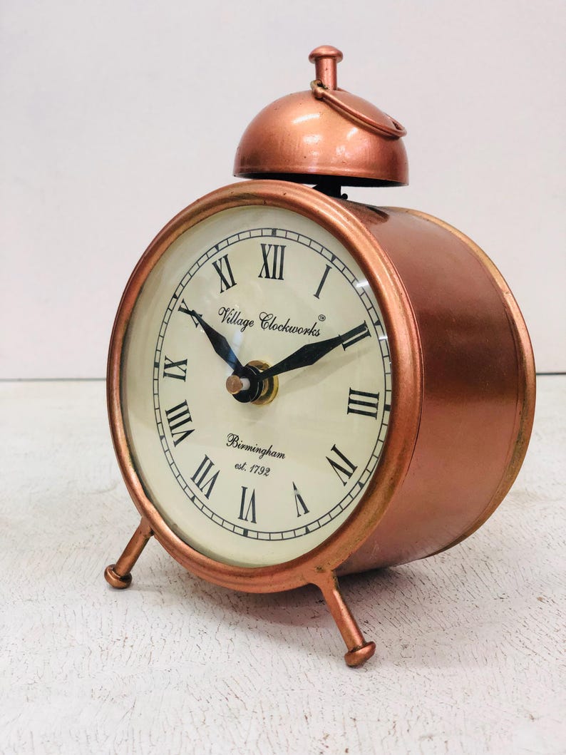 May include: A copper-colored alarm clock with Roman numeral markings on the face. The clock has a bell on top and three legs. The clock face reads "Village Clockworks Birmingham est. 1792".