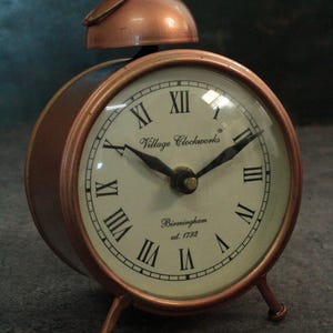 May include: A copper-colored alarm clock with Roman numeral markings on the face. The clock face reads "Village Clockworks Birmingham est. 1792".