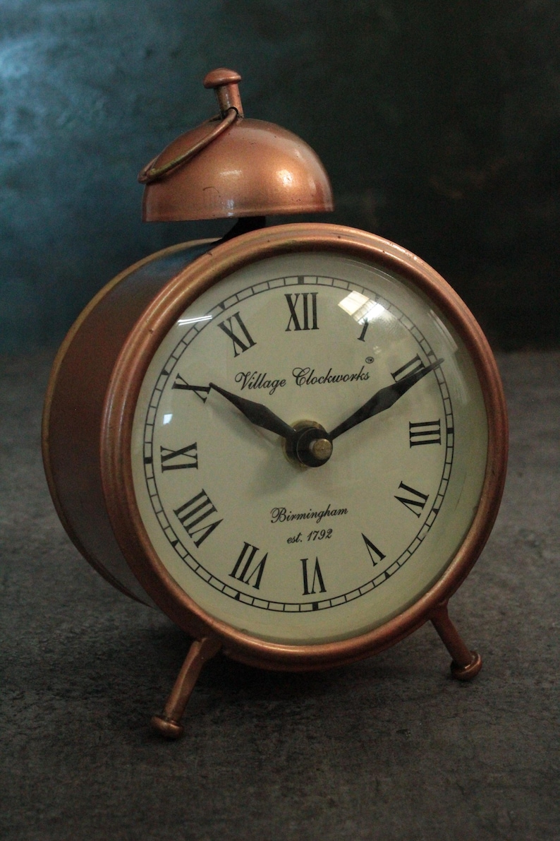 May include: A copper-colored alarm clock with Roman numeral markings on the face. The clock face reads "Village Clockworks Birmingham est. 1792".