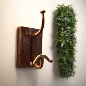Pode incluir: Um gancho de parede rústico com uma base de madeira castanha escura e um gancho de metal curvo e decorativo. O gancho tem um acabamento bronze e é montado numa placa de madeira retangular. À direita, há uma planta verde vertical.