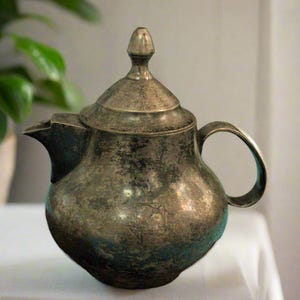 May include: A silver teapot with a tarnished finish and a rounded body. The teapot has a lid with a knob on top and a handle on the side.