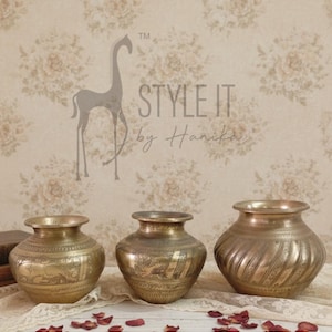 May include: Three antique brass pots with intricate etched designs. The pots vary in size and shape, with one featuring a spiral pattern. The background includes floral wallpaper and the text "STYLE IT by Hanika".