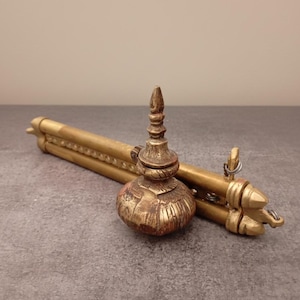 May include: An antique brass-colored incense burner with a decorative, bulbous base and a tall, pointed top. A matching brass-colored tripod is behind the burner, with a series of decorative ridges and a pointed end. The items are on a gray surface.