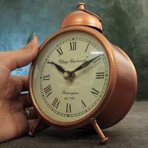 May include: A copper-colored alarm clock with Roman numeral markings on the face. The clock face reads "Village Clockworks Birmingham est. 1792".