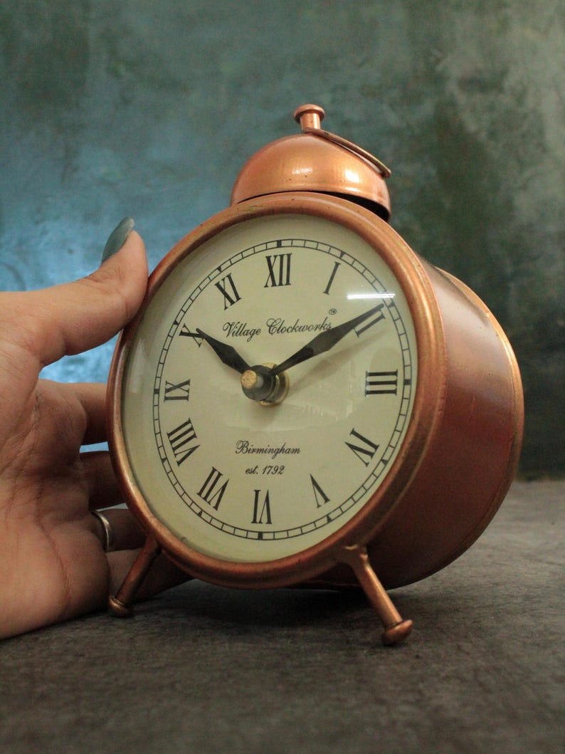 May include: A copper-colored alarm clock with Roman numeral markings on the face. The clock face reads "Village Clockworks Birmingham est. 1792".
