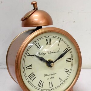 May include: A copper-colored alarm clock with Roman numeral markings on the face. The clock face reads "Village Clockworks Birmingham est 1792".
