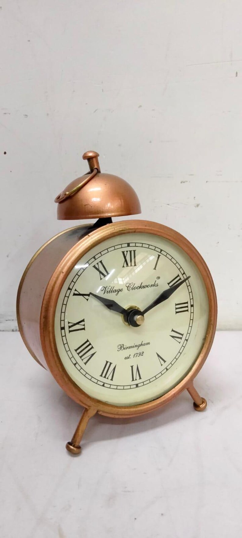 May include: A copper-colored alarm clock with Roman numeral markings on the face. The clock face reads "Village Clockworks Birmingham est 1792".