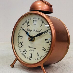 May include: A copper-colored alarm clock with Roman numeral markings on the face. The clock face reads "Village Clockworks Birmingham est. 1792".