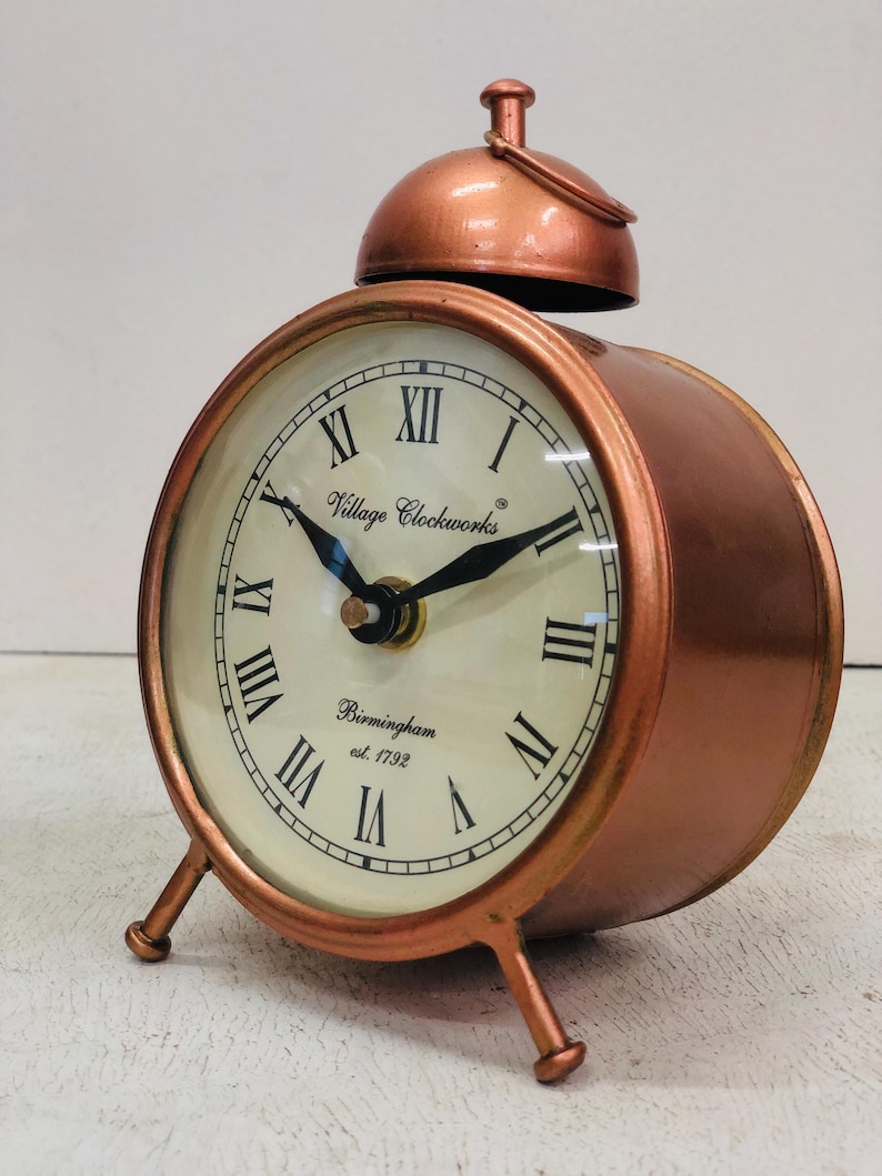 May include: A copper-colored alarm clock with Roman numeral markings on the face. The clock face reads "Village Clockworks Birmingham est. 1792".