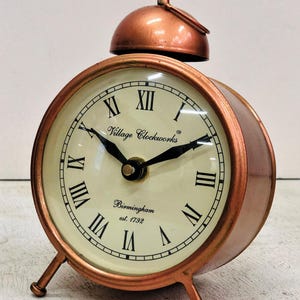 May include: A copper-colored alarm clock with Roman numeral markings on the face. The clock face reads "Village Clockworks" and "Birmingham est. 1792".
