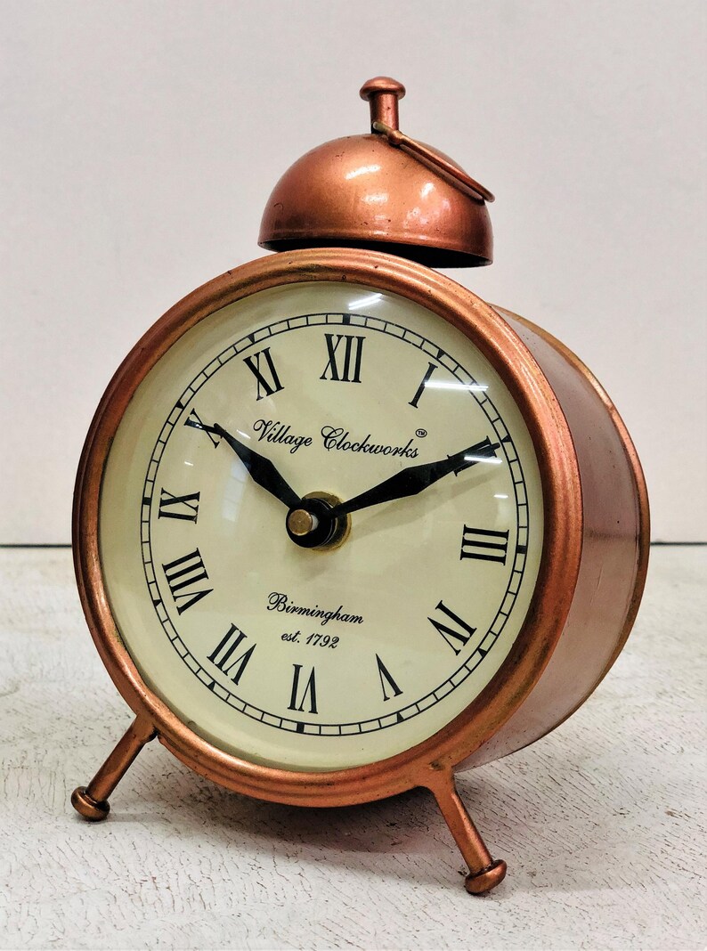 May include: A copper-colored alarm clock with Roman numeral markings on the face. The clock face reads "Village Clockworks" and "Birmingham est. 1792".