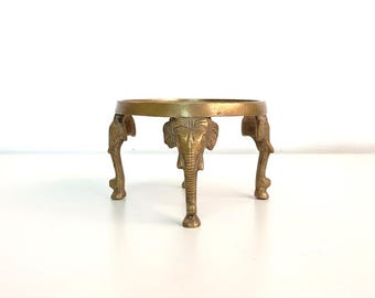 May include: A circular brass stand featuring three legs shaped like elephant heads and trunks. The metal has a warm, metallic gold tone with a smooth finish. The top surface is a flat, open ring design supported by the three distinct elephant figures.