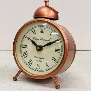 May include: A copper-colored alarm clock with a white face and black Roman numeral hour markers. The clock has a bell on top and three legs. The clock face reads "Village Clockworks Birmingham est. 1792".