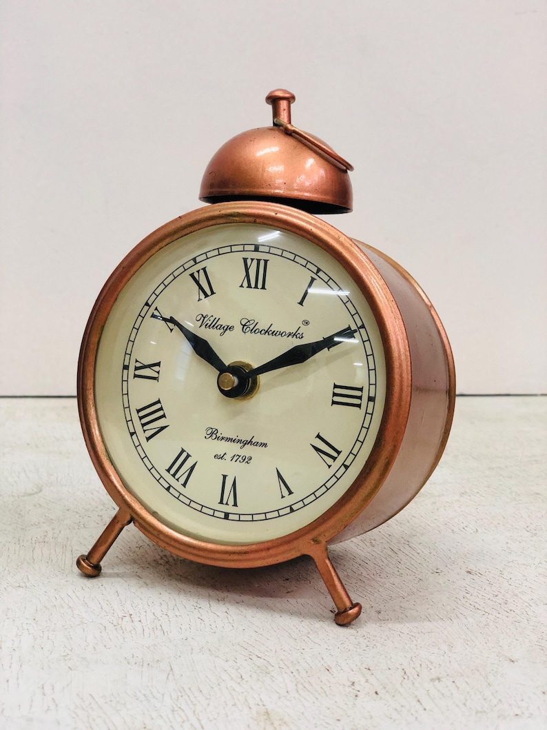 May include: A copper-colored alarm clock with a white face and black Roman numeral hour markers. The clock has a bell on top and three legs. The clock face reads "Village Clockworks Birmingham est. 1792".