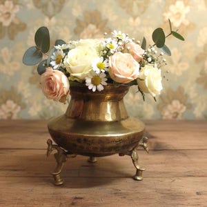 May include: A vintage brass vase with a floral arrangement. The vase has a bulbous body and three decorative feet. The flowers include roses, daisies, and greenery, with soft pink and cream petals. The background features a floral wallpaper.