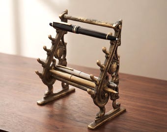 May include: A bronze-toned metal pen display stand featuring an ornate, scroll-like frame with multiple hooks on each side. The stand holds three pens: one black pen resting on the top bar and two metallic pens positioned on the lower hooks.