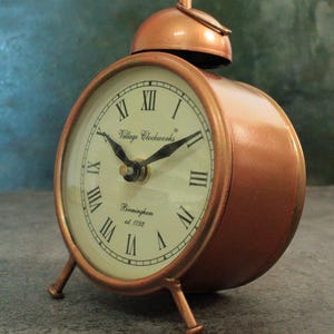 May include: A copper-colored alarm clock with a bell on top. The clock face has Roman numerals and says "Village Clockworks Birmingham est. 1792".