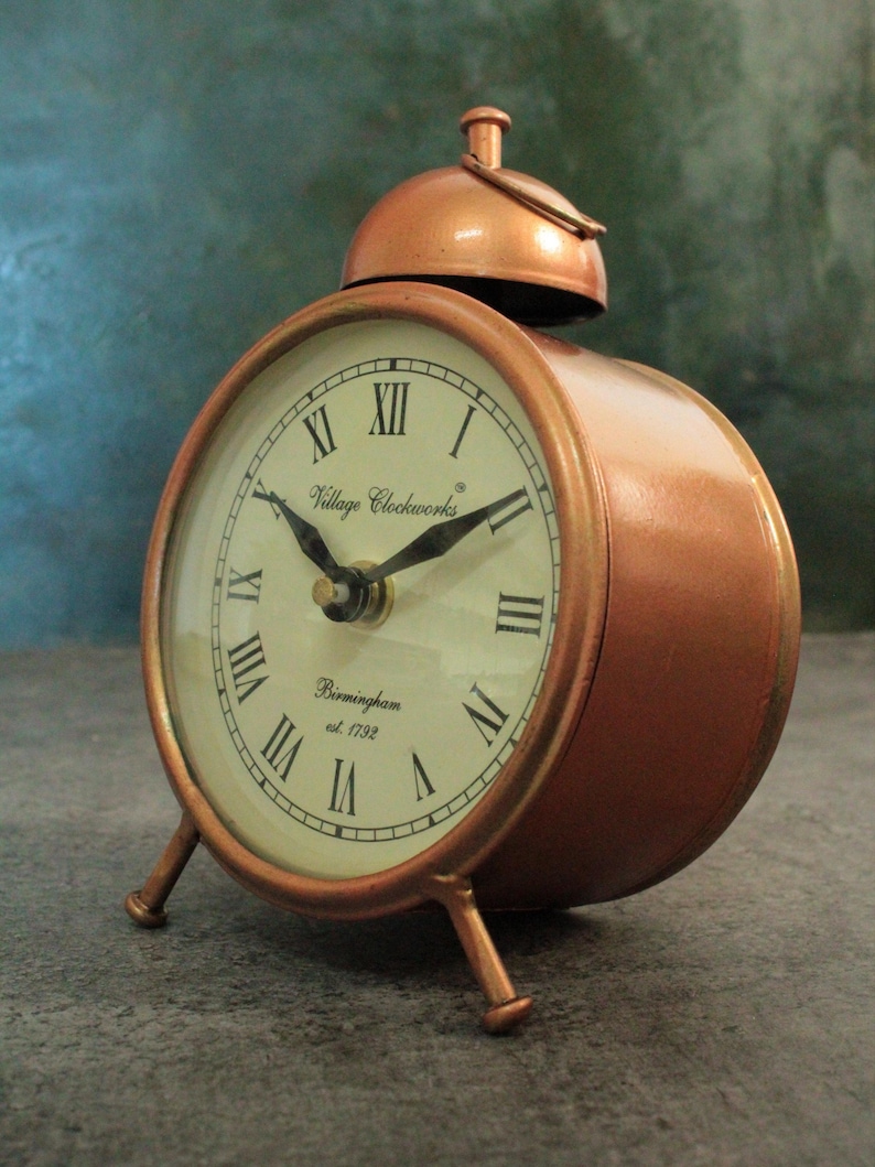 May include: A copper-colored alarm clock with a bell on top. The clock face has Roman numerals and says "Village Clockworks Birmingham est. 1792".