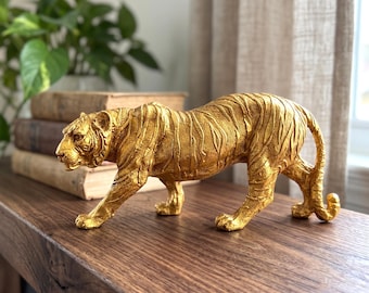 May include: A gold-colored tiger figurine standing in a prowling position. The sculpture features a textured surface that mimics the appearance of fur and distinct tiger stripes across its body, with the head turned slightly to the left side.