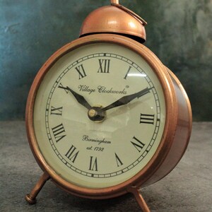 May include: A copper-colored alarm clock with Roman numeral markings on the face. The clock has a bell on top and three legs. The clock face reads "Village Clockworks Birmingham est. 1792".