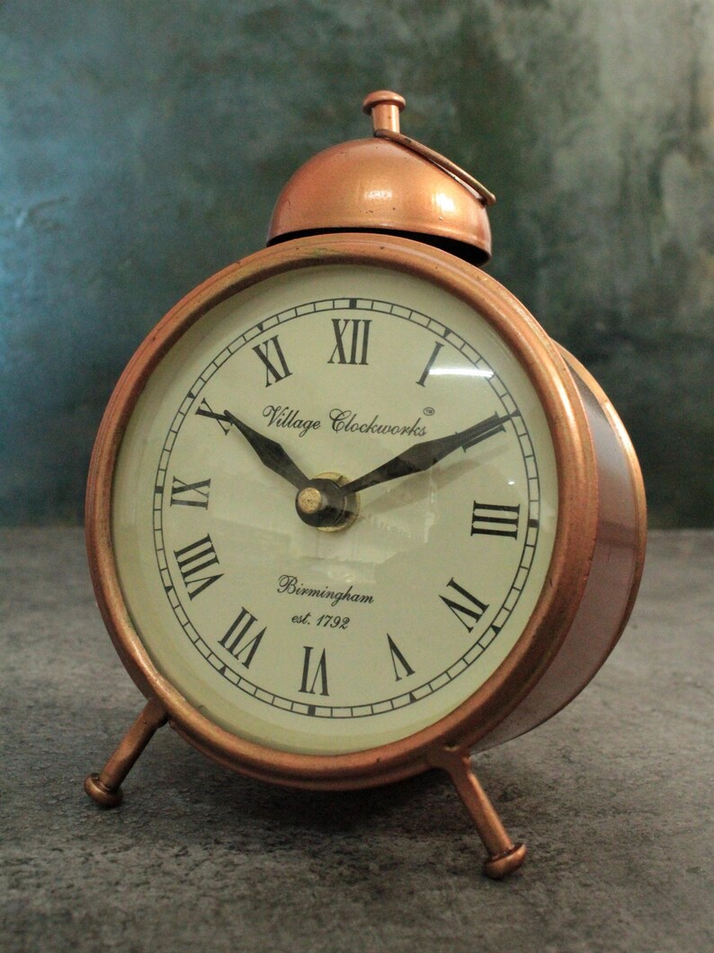 May include: A copper-colored alarm clock with Roman numeral markings on the face. The clock has a bell on top and three legs. The clock face reads "Village Clockworks Birmingham est. 1792".