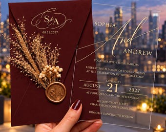 Peut inclure: Une enveloppe bordeaux avec un sceau en cire dorée et des fleurs séchées accompagnée d'une invitation de mariage en acrylique transparent. Le texte doré indique Sophia and Andrew, 21 août 2027, et les détails du Willow Grove Estate.