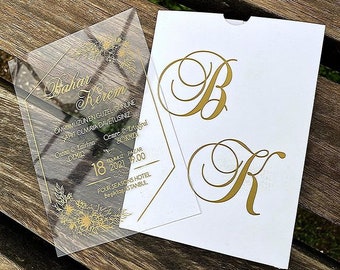May include: A clear acrylic wedding invitation with gold lettering. The invitation features the names "Bahar" and "Kerem" and the date "18 Temmuz 2021". The invitation is decorated with gold floral designs.