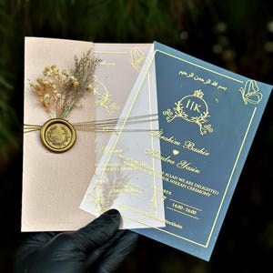Op de afbeelding: Elegante trouwkaarten in poederroze en marineblauw, met goudfolie kalligrafie en decoratieve elementen. De uitnodigingen zijn vastgebonden met een delicate ketting en verzegeld met een gouden wasstempel. De tekst bevat de namen Ibrahim Bashir en Khadra Yasin.