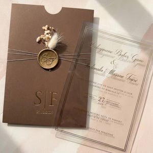Op de afbeelding: Een bruine huwelijksuitnodiging met een transparante overlay. De uitnodiging heeft de initialen "SF" en de datum "20.02.2025". Een gouden waszegel en gedroogde bloemen sieren de voorkant. De transparante overlay heeft tekst.